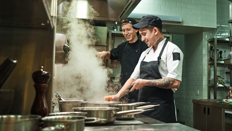 Two chefs in a commercial kitchen with steaming cooking pots