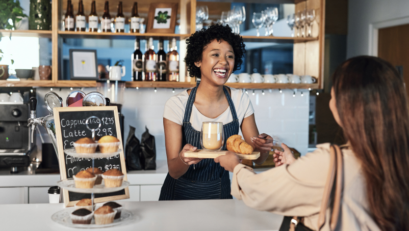 A cafe owner hands a latte and croissant to another woman in a cozy coffee shop setting.