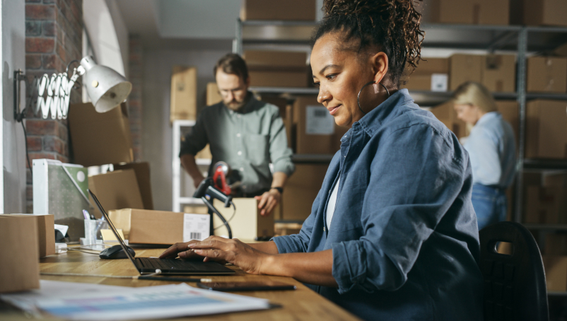 person on a laptop with shipping supplies and coworkers